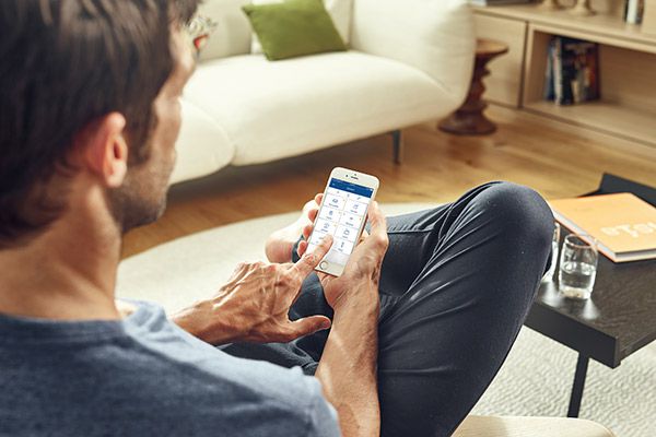 Bildquelle: Gira Ein Mann sitzt auf dem Sofa und bedient mit dem Finger einen Stromsparrechner auf seinem Smartphone im Wohnzimmer.
