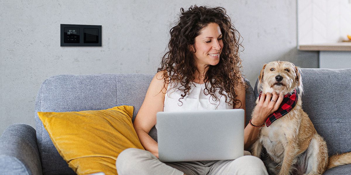 Online-Konfiguratoren von Schneider Electric Frau sitzt mit Laptop und Hund auf Sofa, an der Wand ein modernes Bedienfeld von Schneider Electric.